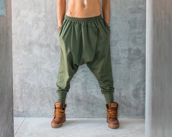 May include: A person wearing olive green harem trousers and brown boots. The person is also wearing a white headband and a beaded necklace. The background is a grey concrete wall.