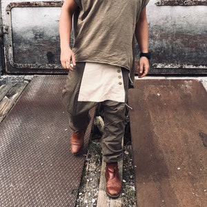 May include: A person wearing a mustard yellow beanie, a loose-fitting olive green t-shirt, and olive green pants with a cream-colored panel. The person is also wearing brown leather boots. The outfit has a casual, bohemian style.