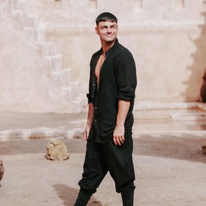 May include: Black linen shirt and pants ensemble. The shirt is buttoned at the collar and open at the chest. The pants are a loose fit. The outfit is paired with black high-top sneakers. The background features a beige building and a sandy ground.