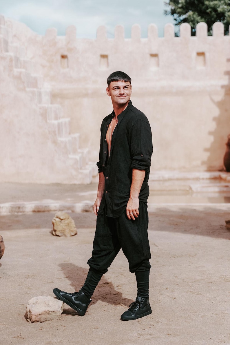 May include: Black linen shirt and pants ensemble. The shirt is buttoned at the collar and open at the chest. The pants are a loose fit. The outfit is paired with black high-top sneakers. The background features a beige building and a sandy ground.