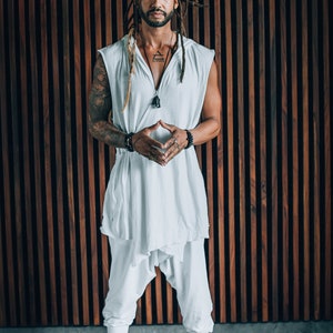 May include: A man with dreadlocks is standing in front of a wood panel wall. He is wearing a white sleeveless shirt with a hood, white trousers, and several bracelets on his wrists. He is looking directly at the camera with his hands clasped in front of him.