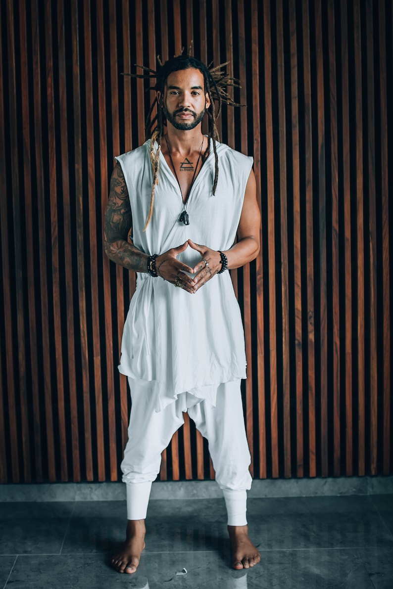 May include: A man with dreadlocks is standing in front of a wood panel wall. He is wearing a white sleeveless shirt with a hood, white trousers, and several bracelets on his wrists. He is looking directly at the camera with his hands clasped in front of him.