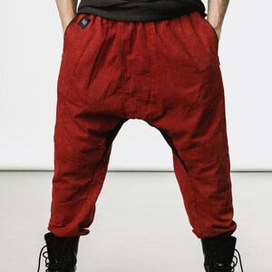 May include: Red drop-crotch pants with a textured appearance. The pants have a relaxed fit, pockets, and stitched details. The model is wearing black boots. The pants are a deep shade of red.