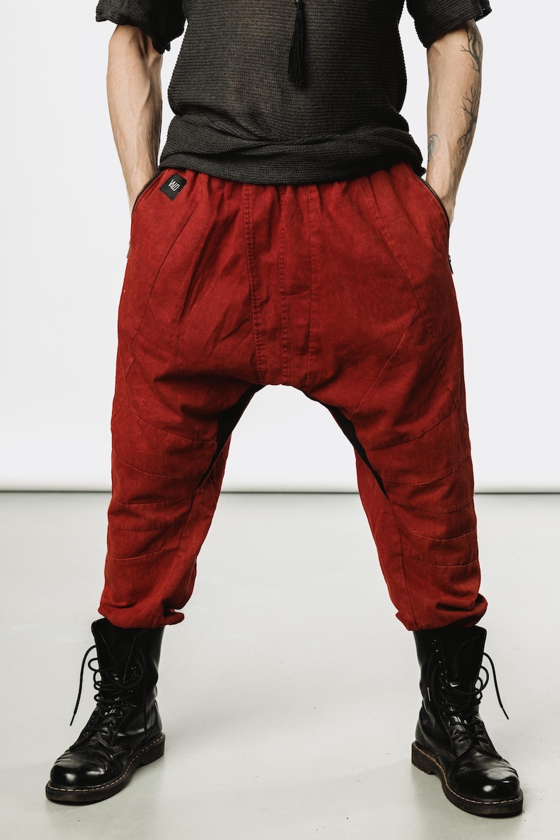 May include: Red drop-crotch pants with a textured appearance. The pants have a relaxed fit, pockets, and stitched details. The model is wearing black boots. The pants are a deep shade of red.