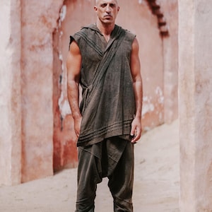May include: A person wearing a dark olive green hooded sleeveless top and matching drop-crotch pants. The outfit has a draped design and is paired with gray ankle cuffs. The background features a textured, reddish-brown wall.