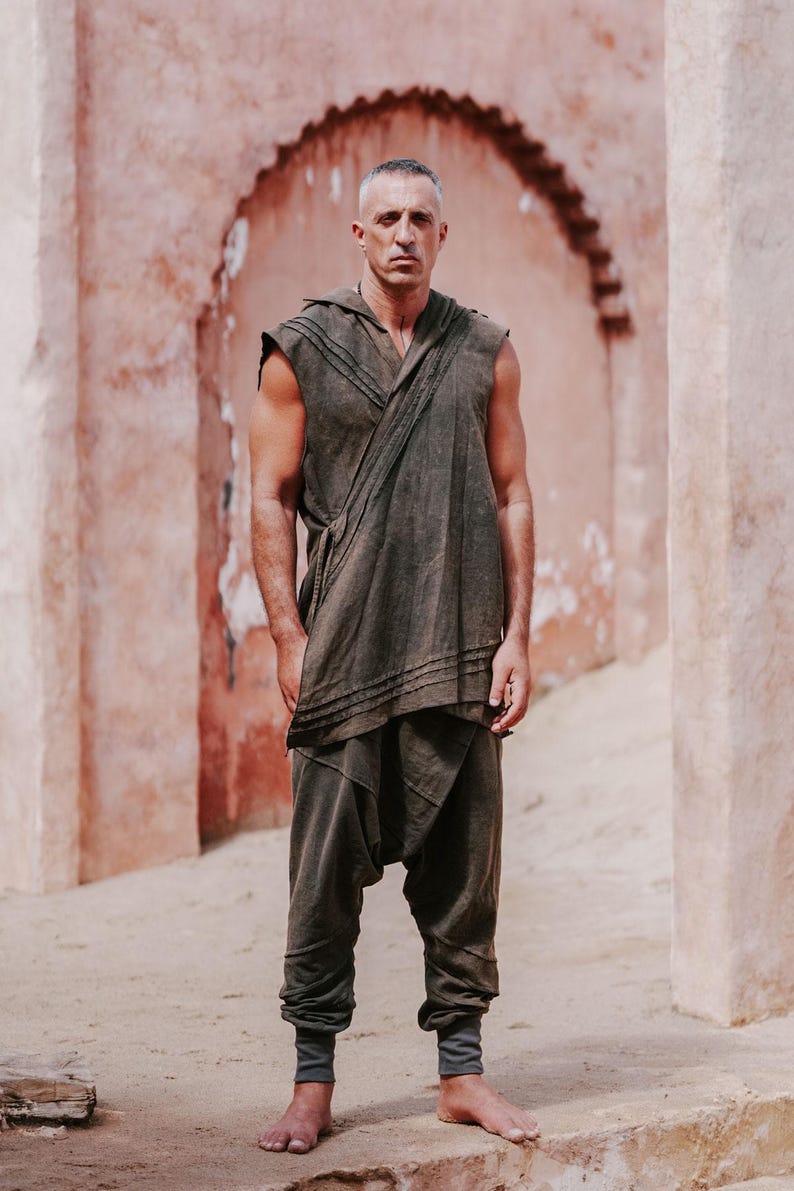 May include: A person wearing a dark olive green hooded sleeveless top and matching drop-crotch pants. The outfit has a draped design and is paired with gray ankle cuffs. The background features a textured, reddish-brown wall.