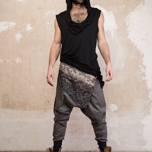 May include: A person wearing a black sleeveless hooded top with a patterned scarf tied around their waist and gray pants. They are wearing brown boots.