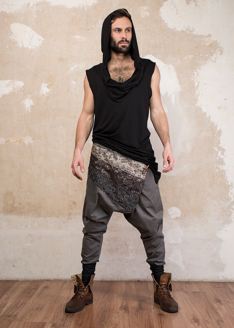 May include: A person wearing a black sleeveless hooded top with a patterned scarf tied around their waist and gray pants. They are wearing brown boots.