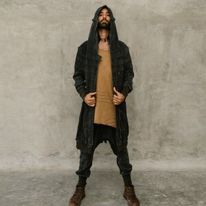 May include: A person wearing a dark gray hooded jacket, a tan shirt, and dark gray pants. The jacket has a long, flowing design with a textured appearance. The pants are a loose fit. The person is also wearing brown boots.