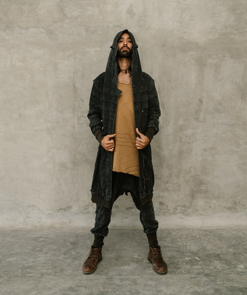 May include: A person wearing a dark gray hooded jacket, a tan shirt, and dark gray pants. The jacket has a long, flowing design with a textured appearance. The pants are a loose fit. The person is also wearing brown boots.