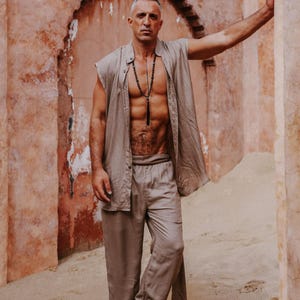 Puede incluir: Un hombre con un conjunto de lino beige, que incluye una camisa sin mangas y pantalones, se encuentra frente a una pared texturizada de color marr&oacute;n rojizo. Lleva un collar largo de cuentas negras. El conjunto parece ligero y adecuado para el clima c&aacute;lido.