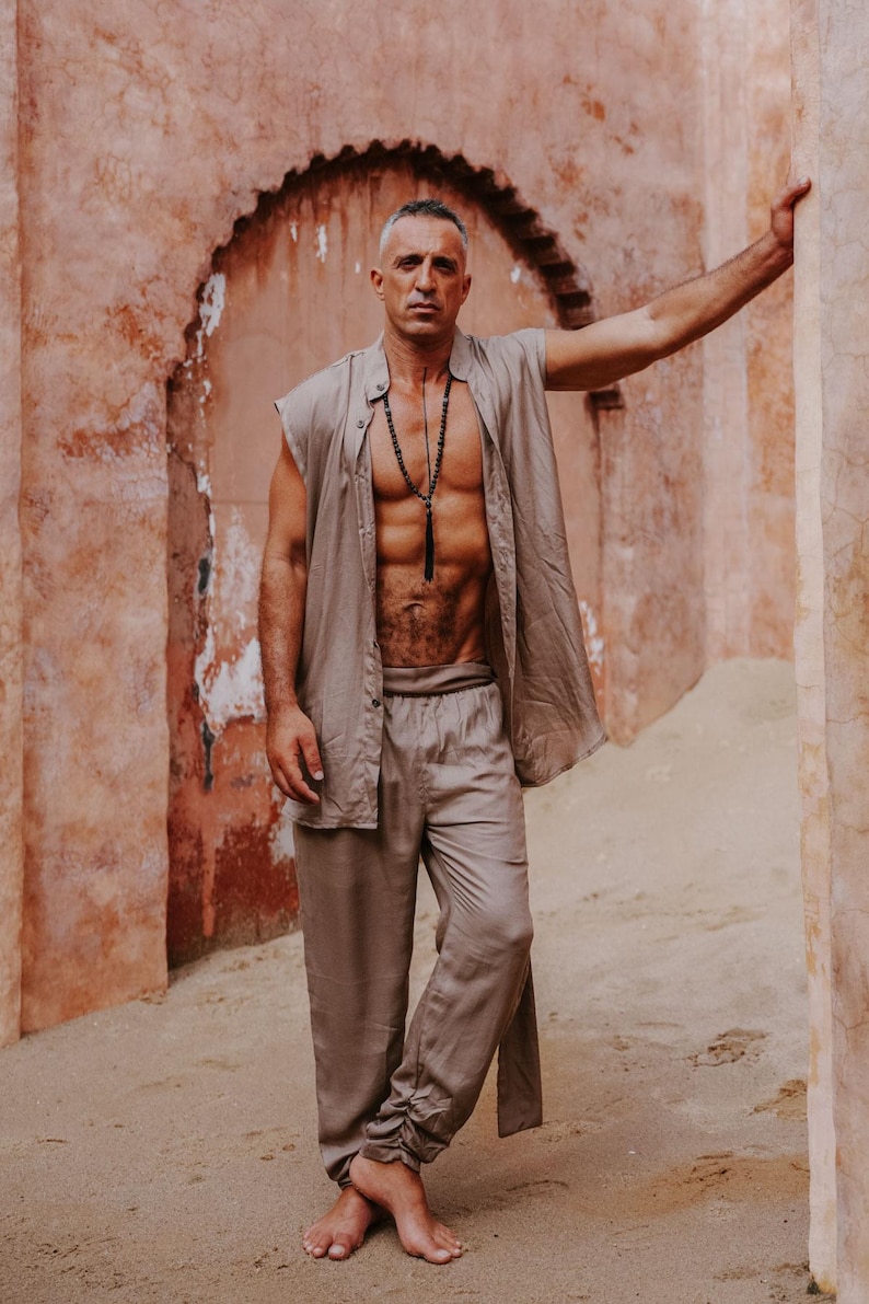 Puede incluir: Un hombre con un conjunto de lino beige, que incluye una camisa sin mangas y pantalones, se encuentra frente a una pared texturizada de color marr&oacute;n rojizo. Lleva un collar largo de cuentas negras. El conjunto parece ligero y adecuado para el clima c&aacute;lido.