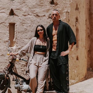 Puede incluir: Dos personas con ropa de lino posan cerca de una motocicleta vintage. Uno viste de negro, el otro de beige. El fondo es una pared clara y texturizada con detalles arquitect&oacute;nicos. Escena soleada.