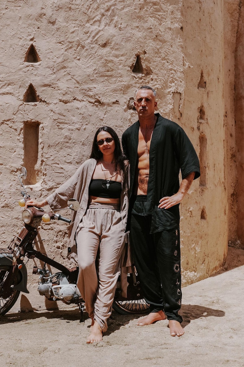 Puede incluir: Dos personas con ropa de lino posan cerca de una motocicleta vintage. Uno viste de negro, el otro de beige. El fondo es una pared clara y texturizada con detalles arquitect&oacute;nicos. Escena soleada.