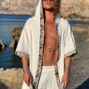 May include: A white hooded robe with brown and white patterned trim. The robe is open in the front and has a silver necklace with a pendant.