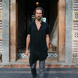 May include: A man wearing a black long-sleeved shirt with a button-down collar and grey pants. He is standing in front of a doorway with a wooden frame and brick walls.