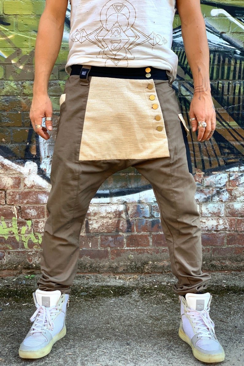 May include: Brown pants with a beige panel secured by gold buttons. The pants have a black waistband and are paired with white high-top sneakers. The person is wearing a white t-shirt with a geometric design.