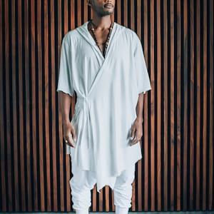 May include: A person wearing a white, loose-fitting robe with a knotted sash and a beaded necklace. The person is standing in front of a wooden wall with vertical slats.