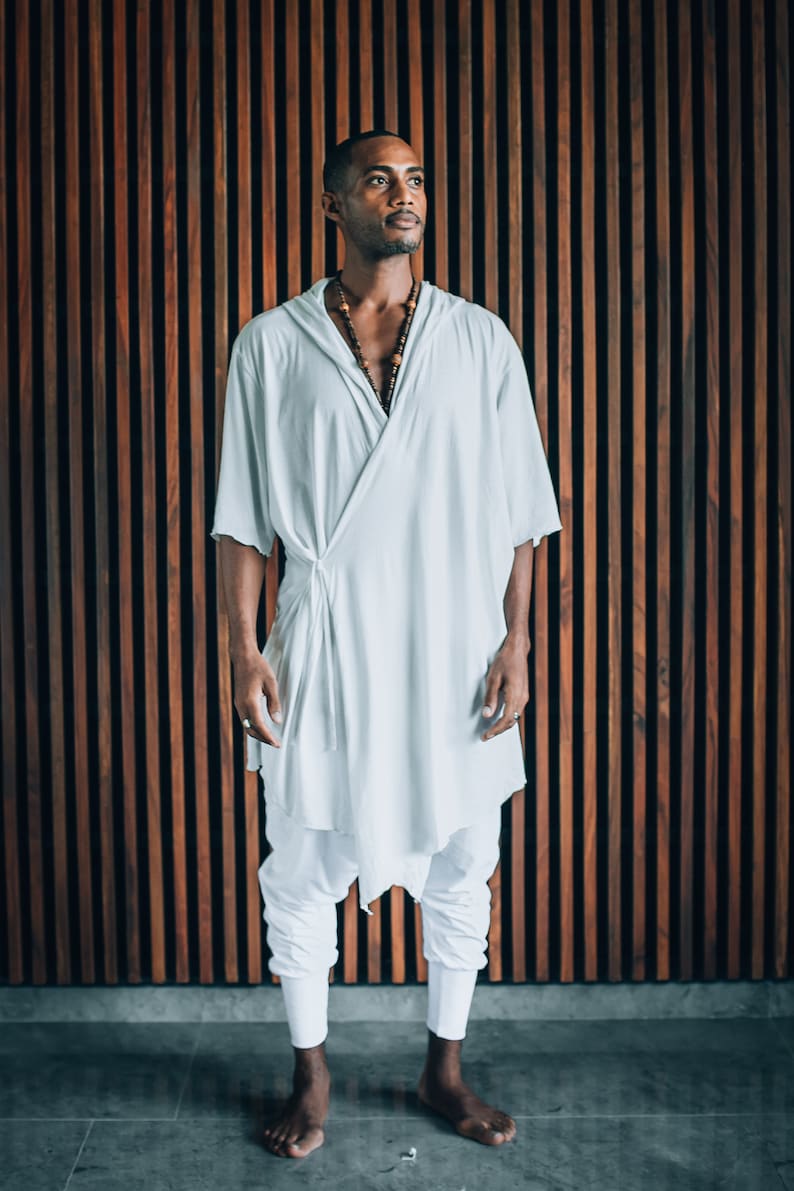 May include: A person wearing a white, loose-fitting robe with a knotted sash and a beaded necklace. The person is standing in front of a wooden wall with vertical slats.
