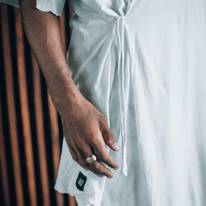 May include: A white, loose-fitting shirt with a long tie that is tied at the waist. The shirt has a simple design and is made of a lightweight fabric. The person wearing the shirt is wearing a gold ring on their finger.