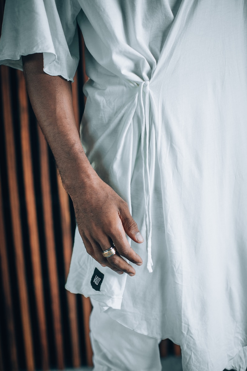 May include: A white, loose-fitting shirt with a long tie that is tied at the waist. The shirt has a simple design and is made of a lightweight fabric. The person wearing the shirt is wearing a gold ring on their finger.