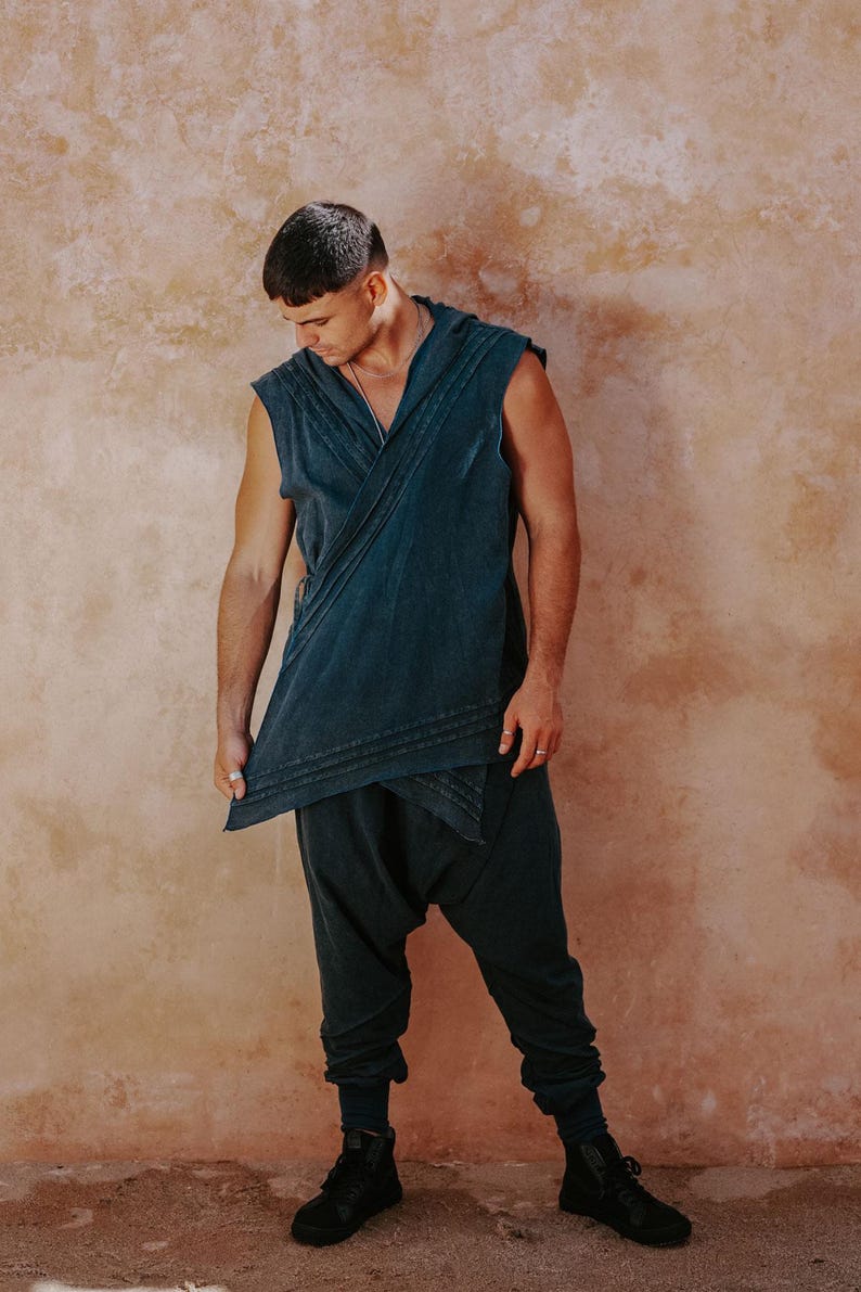 May include: A teal-colored, sleeveless wrap top and matching drop-crotch pants. The top has a V-neck and layered design. The pants have a gathered ankle. The person is wearing black high-top sneakers. The background is a textured, reddish-brown wall.