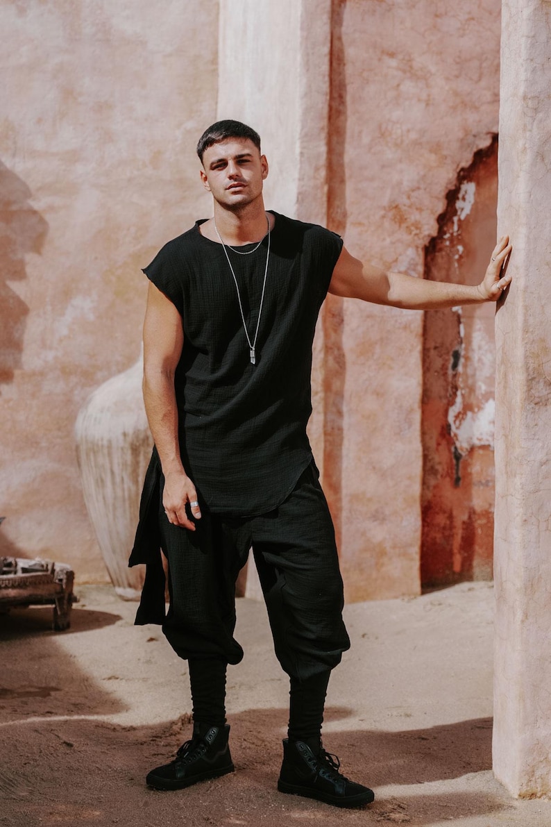 May include: A person wearing a black sleeveless tunic and matching cropped pants. The outfit includes black high-top sneakers and a silver necklace. The background features a textured, peach-colored wall.