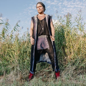 May include: A person wearing a black beanie, black top, and a long vest with a patterned design. The person is also wearing black pants with a purple panel and red boots. The background is a field of tall grass and a blue sky.