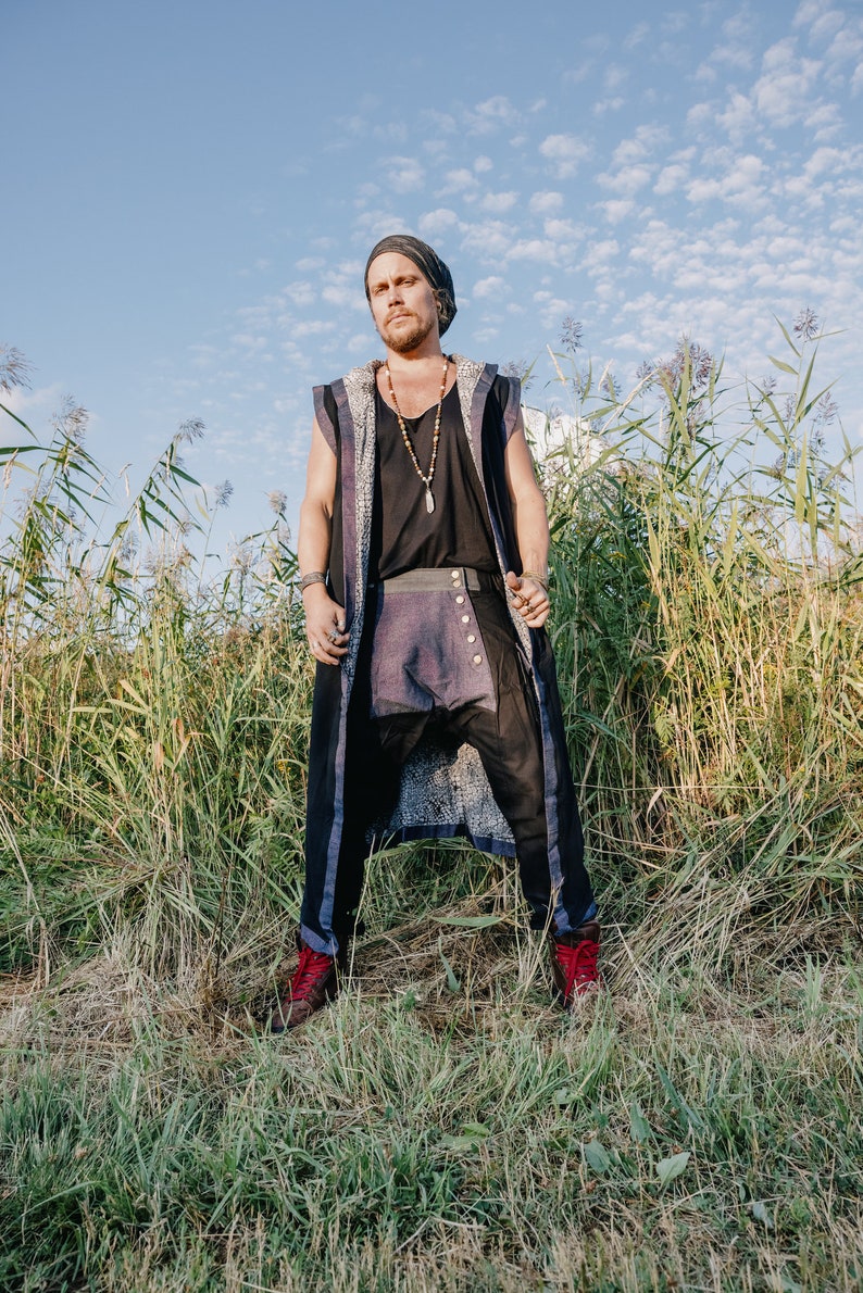 May include: A person wearing a black beanie, black top, and a long vest with a patterned design. The person is also wearing black pants with a purple panel and red boots. The background is a field of tall grass and a blue sky.