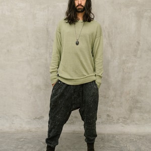 May include: A person wearing a light green sweater, dark gray drop-crotch pants, and brown leather boots. The sweater has a crew neck and long sleeves. The pants have a unique, textured design. A dark pendant hangs from a necklace.