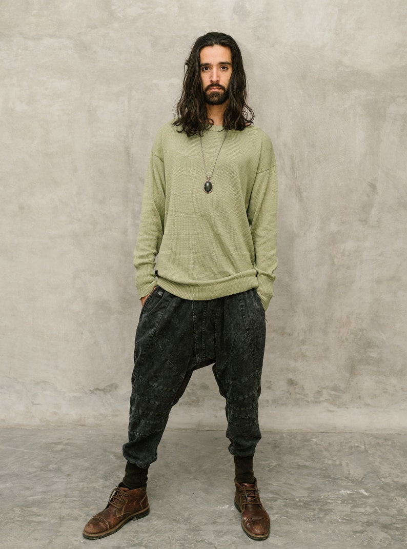 May include: A person wearing a light green sweater, dark gray drop-crotch pants, and brown leather boots. The sweater has a crew neck and long sleeves. The pants have a unique, textured design. A dark pendant hangs from a necklace.