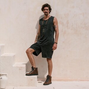 May include: A man wearing a black sleeveless shirt and black shorts with a beaded necklace. He is standing on a white staircase and wearing brown high-top trainers.