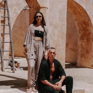 Puede incluir: Una mujer con gafas de sol y top negro est&aacute; de pie junto a un hombre con camisa negra y vaqueros. Est&aacute;n frente a un edificio beige con arcos y una escalera de madera. La mujer lleva un conjunto beige y el hombre est&aacute; sentado.