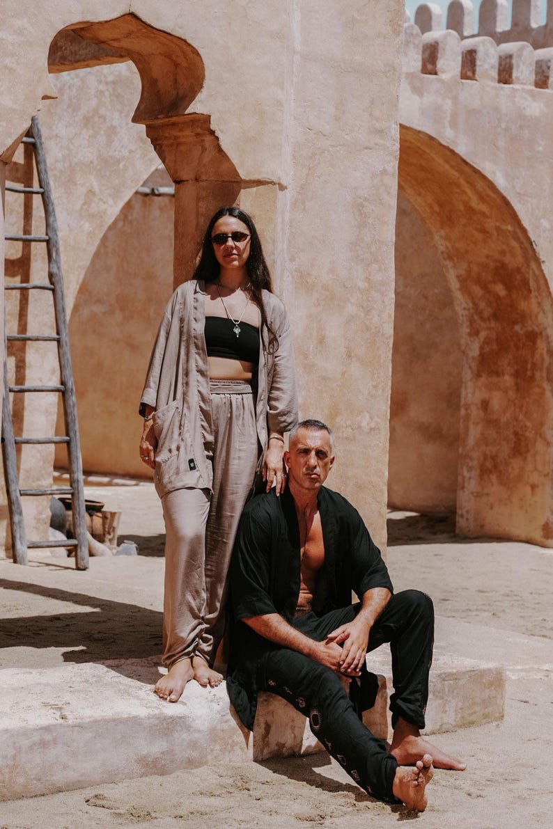 Puede incluir: Una mujer con gafas de sol y top negro est&aacute; de pie junto a un hombre con camisa negra y vaqueros. Est&aacute;n frente a un edificio beige con arcos y una escalera de madera. La mujer lleva un conjunto beige y el hombre est&aacute; sentado.