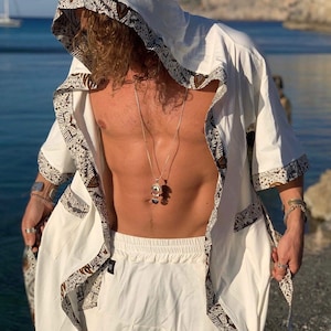 May include: A white hooded robe with brown patterned trim. The robe is open in the front, revealing a white shirt and pants underneath. The person is wearing a silver necklace with a pendant.