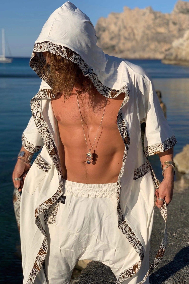 May include: A white hooded robe with brown patterned trim. The robe is open in the front, revealing a white shirt and pants underneath. The person is wearing a silver necklace with a pendant.