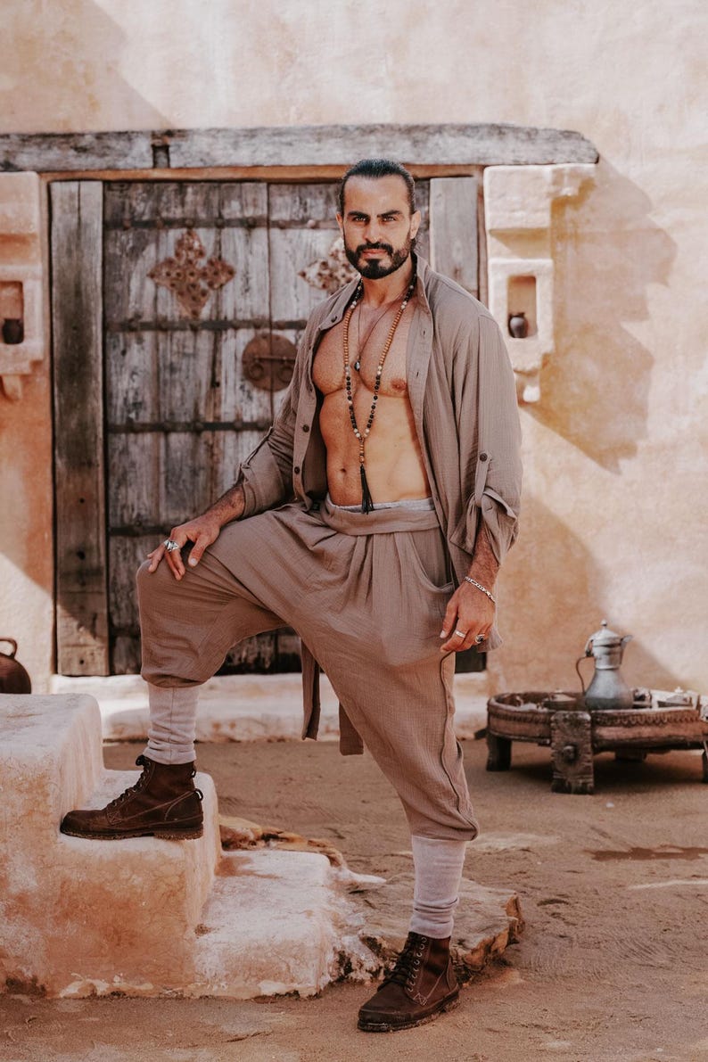 May include: A man in a tan linen outfit, including a button-down shirt and loose pants, stands on stone steps. He wears brown boots and a beaded necklace. The background features a wooden door and a rustic building.