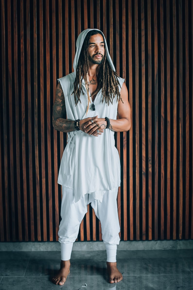 May include: A man with long dreadlocks is wearing a white hooded top with a long, flowing design. He is standing in front of a wood panel wall.