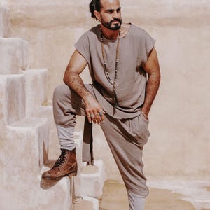May include: A taupe-colored linen outfit consisting of a short-sleeved top and loose-fitting pants. The outfit is paired with brown leather boots. A long beaded necklace adds an accessory. The person is standing on a set of stone steps.