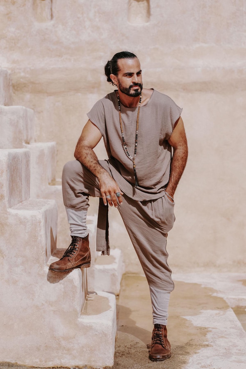 May include: A taupe-colored linen outfit consisting of a short-sleeved top and loose-fitting pants. The outfit is paired with brown leather boots. A long beaded necklace adds an accessory. The person is standing on a set of stone steps.