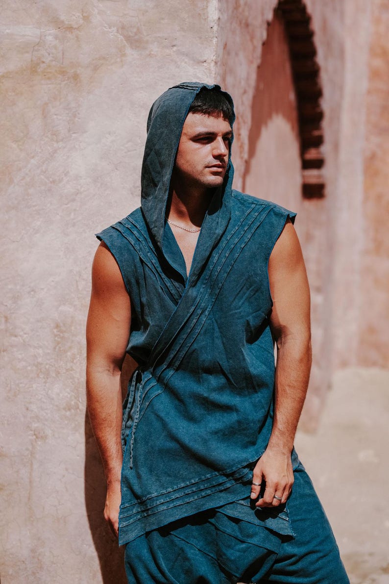 May include: A sleeveless hooded top and matching pants in a faded teal color. The top features a wrap-style design with horizontal stitched detailing. The model is standing in front of a textured, light-colored wall.