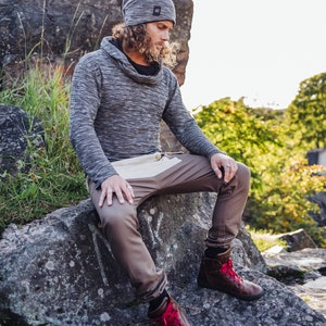 May include: A person wearing a gray knit beanie, a gray cowl-neck sweater, and brown pants with a white panel. They are wearing brown leather boots with red laces. The person is sitting on a large rock outdoors.