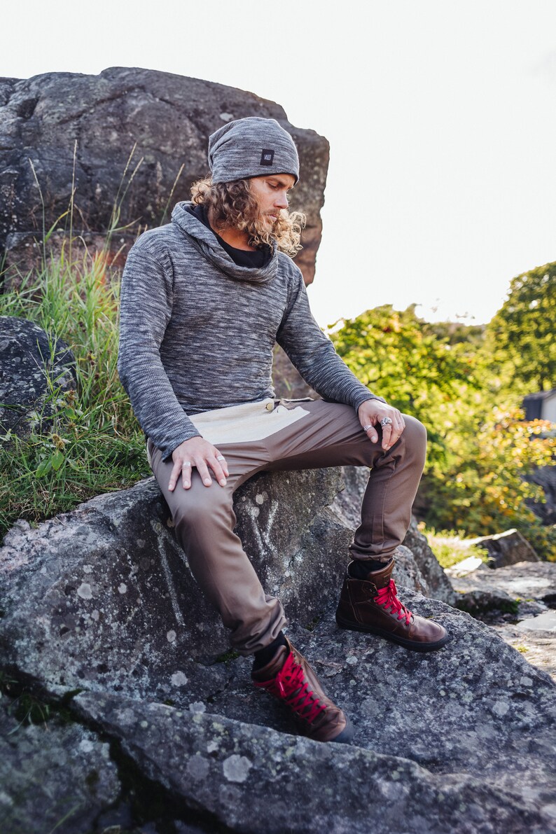 May include: A person wearing a gray knit beanie, a gray cowl-neck sweater, and brown pants with a white panel. They are wearing brown leather boots with red laces. The person is sitting on a large rock outdoors.