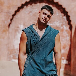 May include: A sleeveless, wrap-style top and matching pants in a faded teal color. The top features a draped design with a hood and tie closure. The model is standing in front of a textured, reddish-brown wall.