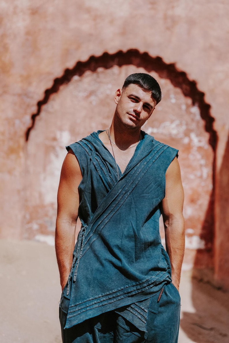May include: A sleeveless, wrap-style top and matching pants in a faded teal color. The top features a draped design with a hood and tie closure. The model is standing in front of a textured, reddish-brown wall.