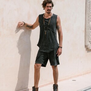 May include: A man wearing a black sleeveless shirt with a distressed look and black shorts. He is wearing a beaded necklace and brown leather high-top trainers.