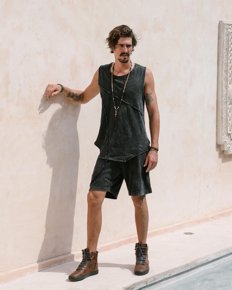 May include: A man wearing a black sleeveless shirt with a distressed look and black shorts. He is wearing a beaded necklace and brown leather high-top trainers.