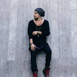 May include: A person wearing a black long-sleeve shirt, black pants, and brown boots with red laces. They are also wearing a gray beanie and a long beaded necklace. The person is leaning against a gray concrete wall.