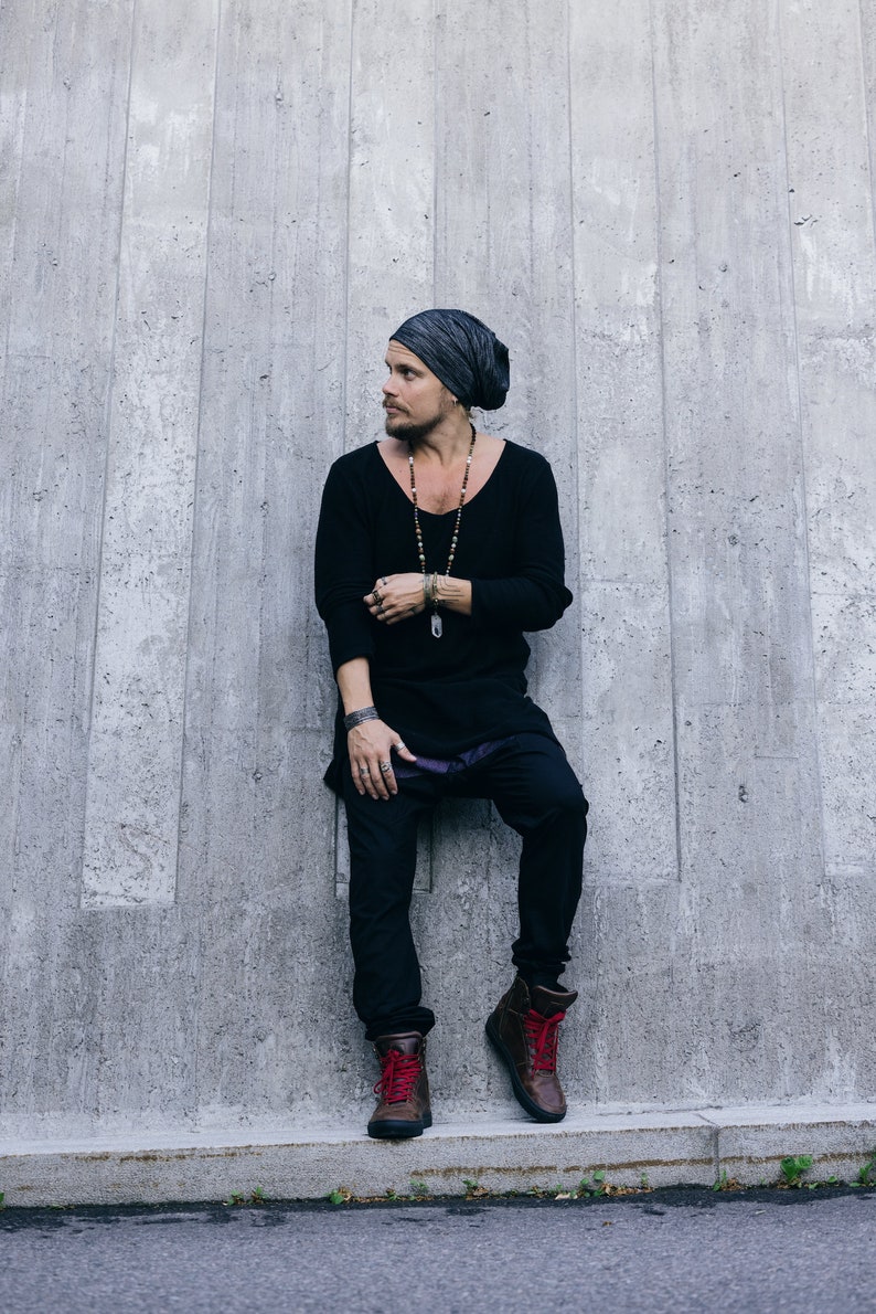 May include: A person wearing a black long-sleeve shirt, black pants, and brown boots with red laces. They are also wearing a gray beanie and a long beaded necklace. The person is leaning against a gray concrete wall.