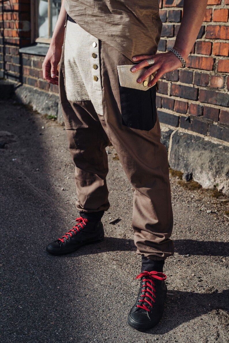 May include: Brown and tan pants with a unique design, featuring a button-up front and a black pocket. The pants are paired with black high-top sneakers with red laces. The outfit is casual and stylish.
